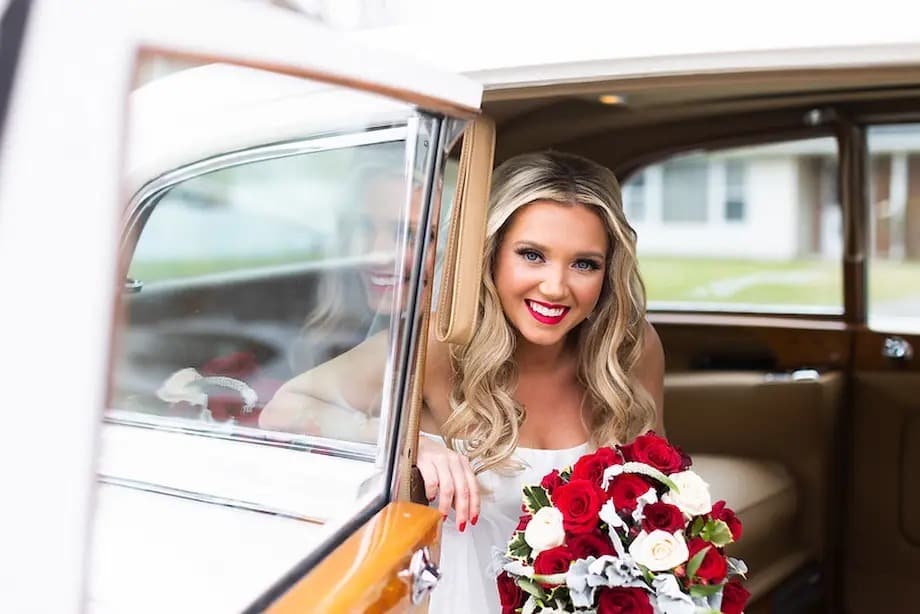 Bride in a wedding dress leaning out of a classic car, holding a red and white rose bouquet